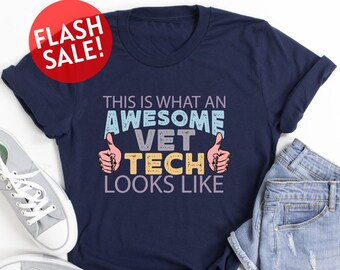 May include: A navy blue t-shirt with the text "This is what an awesome vet tech looks like" in colorful letters with thumbs up on either side. The shirt is laid out on a white surface with a pair of gray canvas shoes and a pair of light blue denim shorts.