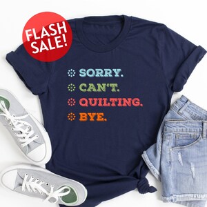 May include: A navy blue t-shirt with the text "SORRY. CAN'T. QUILTING. BYE." in colorful letters. The shirt is laid out on a white surface with a pair of gray sneakers and denim shorts.