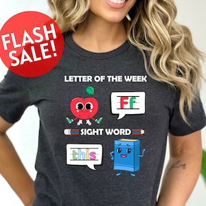 May include: A dark grey t-shirt with a graphic design featuring a red apple, a blue book, and a pencil. The text "Letter of the Week" and "Sight Word" are also included in the design.