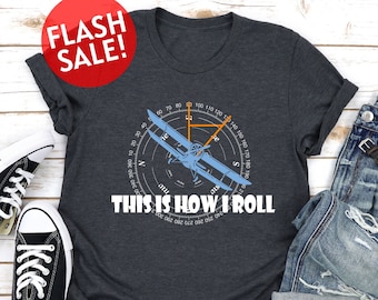 Może przedstawiać: Szary t-shirt z grafiką kompasu i samolotu. Pod grafiką nadrukowany jest tekst "THIS IS HOW I ROLL".