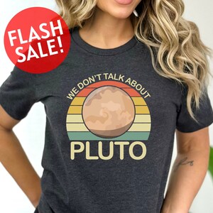 May include: A dark grey t-shirt with a retro-style graphic of Pluto. The graphic features a sunset with the text "We don't talk about Pluto".