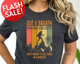 May include: A dark gray t-shirt with a vintage-style graphic design. The graphic features a hiker on a mountain with the text "Out of Breath Hiking Society" and "Don't worry I'll be there in a minutes".