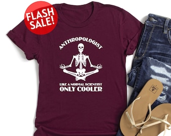 May include: A maroon t-shirt with a white skeleton sitting in a yoga pose. The text on the shirt reads "Anthropologist Like a Normal Scientist Only Cooler".