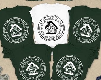 May include: A flat lay featuring a white t-shirt and four forest green t-shirts, all with a circular graphic of a gingerbread house and the text "Gingerbread House Decorating Crew". Additional items include sunglasses, a clock, and jeans.