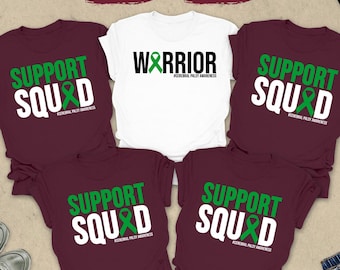 May include: A collection of maroon and white t-shirts with the words "SUPPORT SQUAD" and "WARRIOR" printed on them in green and white. The shirts are arranged with accessories like sunglasses, a clock, and jeans.