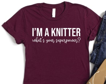 May include: A maroon t-shirt with white text that reads "I'm a knitter what's your superpower?" The shirt is laid flat on a white background with a pair of blue jeans and brown sandals beside it.