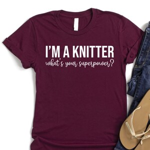May include: A maroon t-shirt with white text that reads "I'm a knitter what's your superpower?" The shirt is laid flat on a white background with a pair of blue jeans and brown sandals beside it.