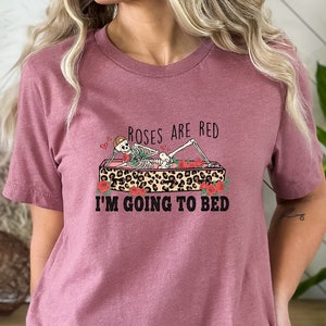 May include: A pink t-shirt with a skeleton lying in a coffin with roses and the text "Roses are red, I'm going to bed".