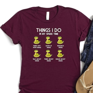 May include: A maroon t-shirt with a white graphic that says "Things I do in my spare time" and features six cartoon snakes with text below each snake that says "Hang out with snake", "Look at snake", "Research snake", "Talk about snake", "Think about snake", and "Dream about snake".