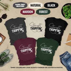 a collection of six t-shirts with the same design, arranged on a beige background. The t-shirts feature the text "Straight Tripin'" in a stylized font. The t-shirts are in various colors, including dark grey, maroon, natural, black, forest, and maroon. There are also sunglasses, a clock, a pen, a plant, and a pair of jeans in the scene. The text "SHIRT COLORS MAY LOOK SLICTER OR Lighter IN PHOTOS" is also present, indicating that the colors of the t-shirts may appear differently in photographs.