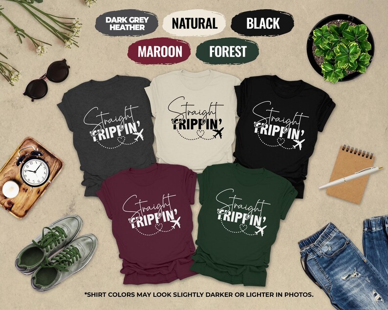 a collection of six t-shirts with the same design, arranged on a beige background. The t-shirts feature the text "Straight Tripin'" in a stylized font. The t-shirts are in various colors, including dark grey, maroon, natural, black, forest, and maroon. There are also sunglasses, a clock, a pen, a plant, and a pair of jeans in the scene. The text "SHIRT COLORS MAY LOOK SLICTER OR Lighter IN PHOTOS" is also present, indicating that the colors of the t-shirts may appear differently in photographs.