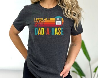 May include: A dark grey t-shirt with a retro-style graphic of a floppy disk and the text "I keep all my dad jokes in a dad-a-base".