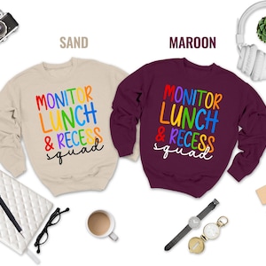 May include: Two crewneck sweatshirts, one sand-colored and one maroon, are displayed. Both feature the text "MONITOR LUNCH & RECESS squad" in colorful, rainbow-style lettering. Various accessories, including a camera, headphones, and a watch, surround the sweatshirts.