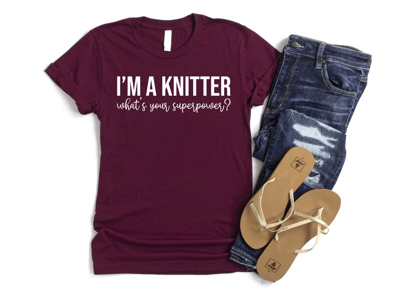 May include: A maroon t-shirt with white text that reads "I'm a knitter what's your superpower?" The shirt is laid flat on a white background with a pair of blue jeans and brown sandals beside it.