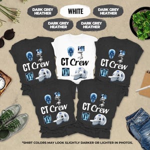 May include: A display of CT Crew t-shirts in white and dark grey heather. Each shirt features a CT scan image, a skull, and the words "CT Crew". The shirts are arranged with accessories like sunglasses, a clock, and jeans.