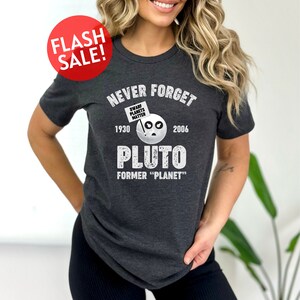 May include: A dark gray t-shirt with a white graphic that says "Never Forget Pluto" with the years 1930 and 2006 on either side of the word Pluto. The graphic also includes a cartoon of Pluto with a sign that says "Dwarf Planets Matter".