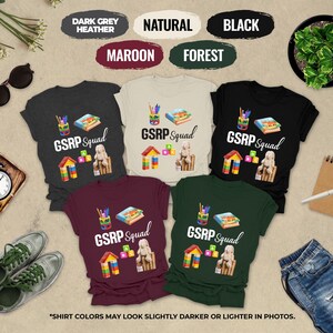 May include: A collection of t-shirts in various colors, including dark grey heather, natural, black, maroon, and forest green. Each shirt features the text "GSRP Squad" along with graphics of school supplies and building blocks. The shirts are displayed with accessories.