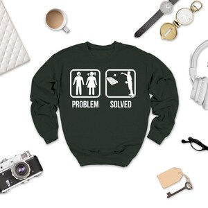 May include: A dark green crewneck sweatshirt featuring a white graphic print. The design displays two square icons: the first shows a person standing next to another person with the text "PROBLEM" below, and the second shows a person throwing a beanbag at a cornhole board with the text "SOLVED" below.