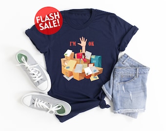 May include: A navy blue t-shirt with a graphic of a hand sticking out of a pile of boxes and mail. The text "I'M OK" is printed on the shirt.