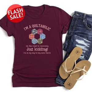 May include: Burgundy t-shirt with a white graphic that says "I'm a Quiltaholic on the road to recovery Just kidding! I'm on my way to buy more fabric". The graphic also features a colorful hexagon design.