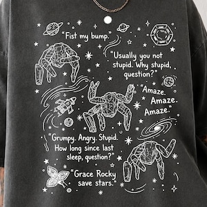 May include: A charcoal grey oversized t-shirt featuring a white graphic print of space-themed illustrations, including planets, stars, and rock-like creatures. The text reads: Fist my bump, Usually you not stupid, Why stupid question, Amaze, Grumpy, Angry, Stupid, How long since last sleep, question, Grace Rocky save stars.