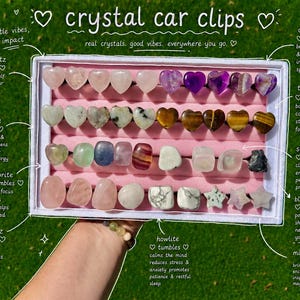 May include: A white rectangular display box containing an assortment of crystal car clips arranged in rows on a pink velvet insert. The collection includes heart-shaped, tumbled, and star-shaped stones in various colors, labeled with text.