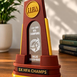 May include: A maroon and gold trophy featuring a central clear panel with the text 2004-2005 HFA CHAMPIONS, DALLAS GOLDEN NUKES, and a logo. The base includes a black plaque with the text 5X HFA CHAMPS, and a gold circular emblem at the top with HFA.
