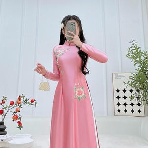 May include: A long, pink dress with long sleeves and a mandarin collar. The dress features floral embroidery on the chest and side, with a white stripe detail. The website address AodaiEmily.com is visible.