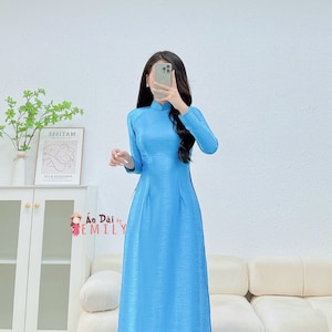 May include: A light blue long-sleeved dress with a mandarin collar and side slits. The dress is made of a silky fabric and has a subtle sheen. The dress is photographed against a white wall and a white sofa.