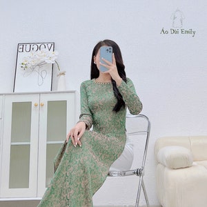 May include: A green and beige patterned long-sleeved dress with a beaded neckline. The dress is worn by a person sitting on a chair. The text "Ao Dai Emily" is visible in the image.