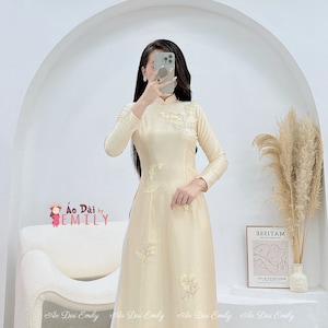 May include: A cream-coloured silk áo dài with delicate floral embroidery. The áo dài is a traditional Vietnamese garment.