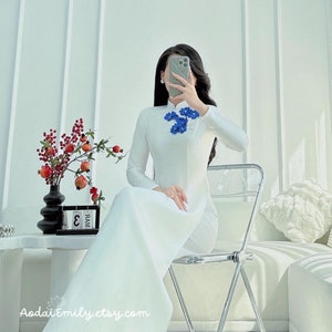 Könnte beinhalten: Ein weißes, langärmeliges Kleid mit Stehkragen und blauer Blumenstickerei. Das Kleid wird von einer Person getragen, die auf einem Stuhl sitzt. Das Kleid ist lang und fließend, und die Person trägt weiße Schuhe.