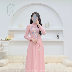 May include: A pink traditional Vietnamese dress with long sleeves and a floral embroidery design. The dress has a high collar and a long skirt. The dress is photographed in front of a white wall with a white sofa in the foreground.
