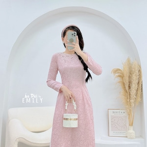 Puede incluir: Un vestido rosa de manga larga con ribete blanco y escote de cuentas. El vestido lo lleva una persona que sostiene un bolso blanco. El texto "Áo Dài by EMILY" es visible en la imagen.