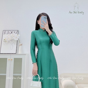 May include: A green silk dress with a white trim and pearl accents. The dress has a mandarin collar and long sleeves. The dress is being held by a person wearing a white handbag.