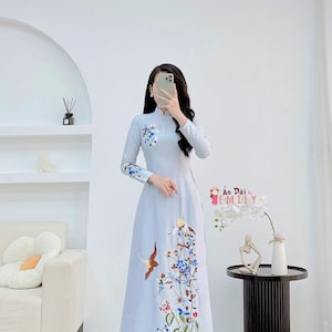May include: A light blue long-sleeved dress with a floral and bird embroidery design. The dress has a mandarin collar and a long skirt. The text "Ao Dai by EMILY" is visible in the image.