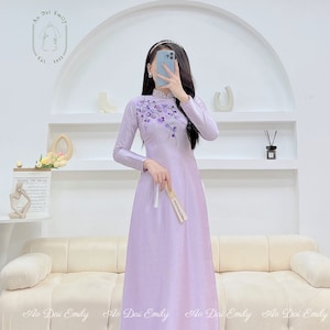 May include: A lavender silk dress with a floral embroidery design. The dress has long sleeves and a mandarin collar. The dress is a traditional Vietnamese garment called an "Ao Dai".