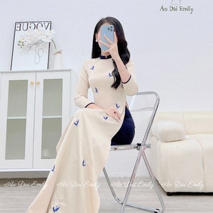 May include: A beige long-sleeved dress with a high collar and a side slit. The dress has a delicate floral embroidery pattern with blue birds. The dress is worn by a person sitting on a chair. The text "Ao Dai Emily" is visible in the image.