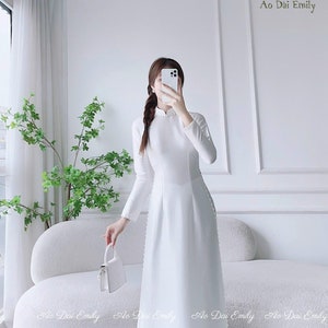 May include: A white long-sleeved dress with a mandarin collar and pearl detailing. The dress is worn by a person holding a white handbag and a white phone. The background is a white wall with a white couch.