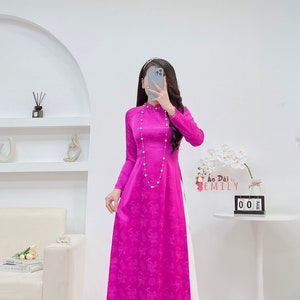 May include: A long, pink, silk dress with a white panel down the side. The dress has a mandarin collar and a floral pattern. The dress is worn by a person with long black hair. The text "Ao Dai by Emily" is visible on the dress.