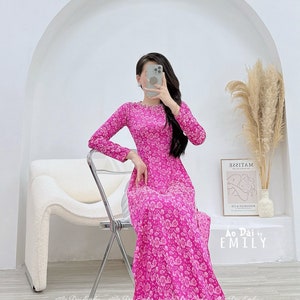 May include: A pink long-sleeved dress with a floral print. The dress has a round neckline and a long skirt. The dress is made of a soft, flowing fabric. The dress is photographed on a model standing in front of a white wall. The text "Ao Dai by EMILY" is visible in the image.