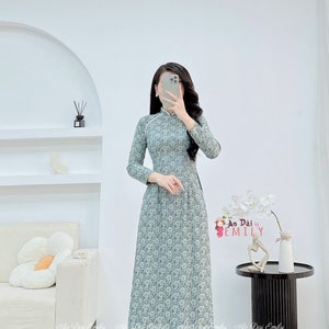 May include: A long-sleeved, floor-length dress in a light blue floral pattern. The dress features a mandarin collar and a fitted bodice that flows into a flared skirt. The text "Ao Dai Emily" is visible in the image.