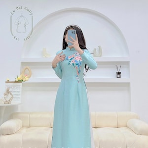 May include: A light blue traditional Vietnamese dress with a white floral design on the front. The dress has long sleeves and a high neckline. The dress is called an "Ao Dai" and is a popular choice for special occasions.