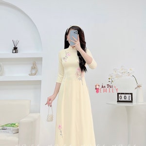 以下が含まれることがあります： 花柄の刺繍が施された薄い黄色のベトナムの伝統的なドレス。ドレスは、マンダリンカラーと七分袖です。ドレスは白いパンツと白い靴と組み合わせています。画像には「Ao Dai by Emily」というテキストが表示されています。