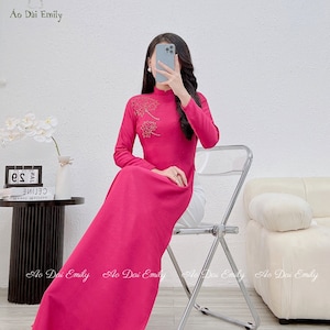 May include: A pink long-sleeved dress with a high collar and a floral design on the chest. The dress is being worn by a person sitting on a chair. The text "Ao Dai Emily" is visible in the top left corner of the image.