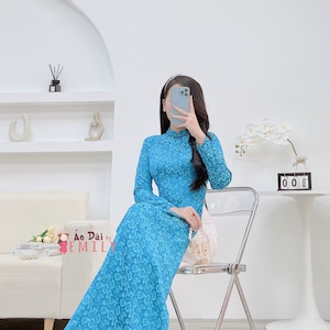 May include: A woman wearing a blue and white floral print long-sleeved dress with a high neck. The dress has a side slit and is sitting on a silver folding chair. The background is a white wall with a shelf and a white orchid plant.