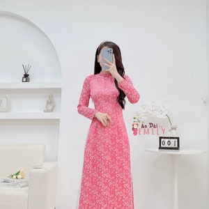 May include: A pink long-sleeved dress with a white floral pattern and a white trim. The dress has a high collar and a side slit. The dress is called "Ao Dai by Emily".