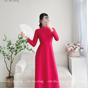 May include: A vibrant pink traditional Vietnamese dress, known as an Ao Dai, with a high collar and long sleeves. The dress is adorned with a row of white pearls down the front and along the sleeves. The dress is worn by a person standing in front of a white sofa and a potted plant.