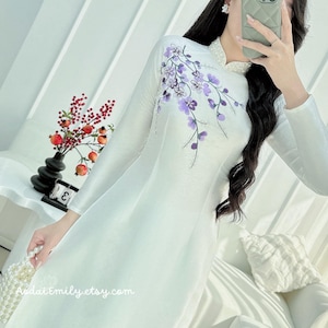 Puede incluir: Vestido blanco de seda con mangas largas y cuello mandarín. El vestido presenta un diseño floral morado en el lado derecho. El vestido se muestra en una modelo con cabello negro largo.
