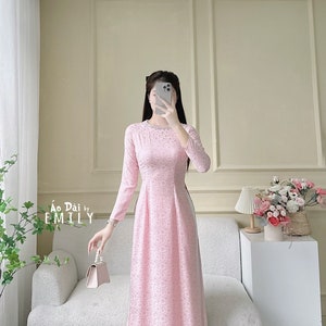 May include: A pink long-sleeved dress with a white trim and floral pattern. The dress has a high neckline and a side slit. The dress is being held by a person with a small white purse.